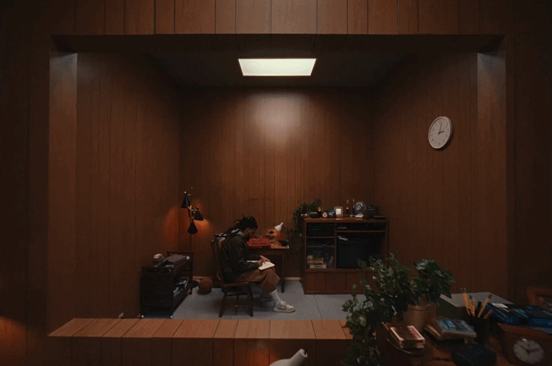 a gif from a J. Cole music video directed by Ryan Doubiago, depicting a moving shot looping through different eras of the rapper's life, all taking place in the same studio walls. in this shot, the musician is working on creating music in the studio.