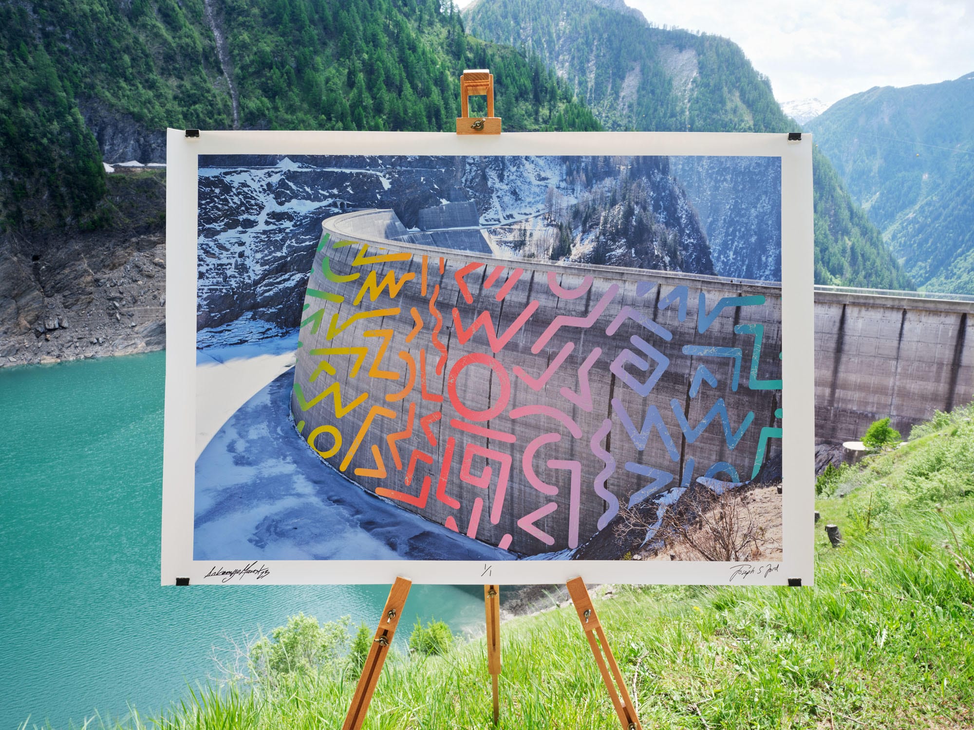 A photographic print of a dam in Switzerland with a street art intervention superimposed on the image, which is photographed on an easel in front of the same landscape