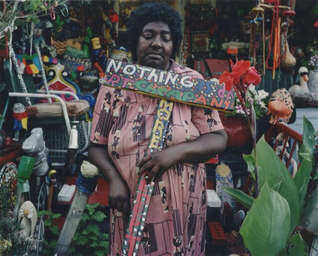 A photograph of artist L.V. Hull holding a hand-painted wooden sign that reads "nothing is dirt cheap anymore" in her garden, outside of her artful home