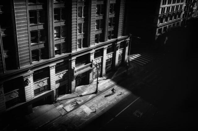a black-and-white photograph by Daniel Sackheim in a noir style of an elevated city street view with a figure walking amid deep shadows