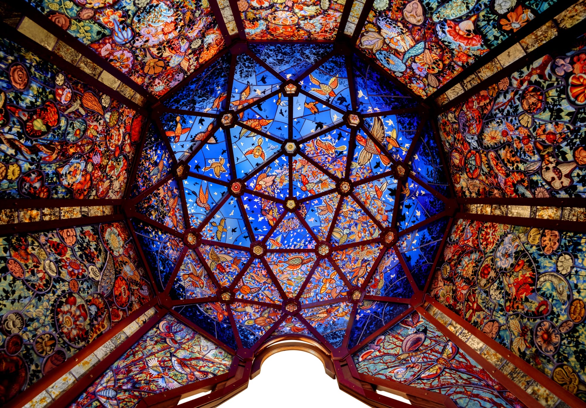 A view from inside a domed, stained glass sculpture by Judith Schaechter, looking up at images of flowers, birds, and other images