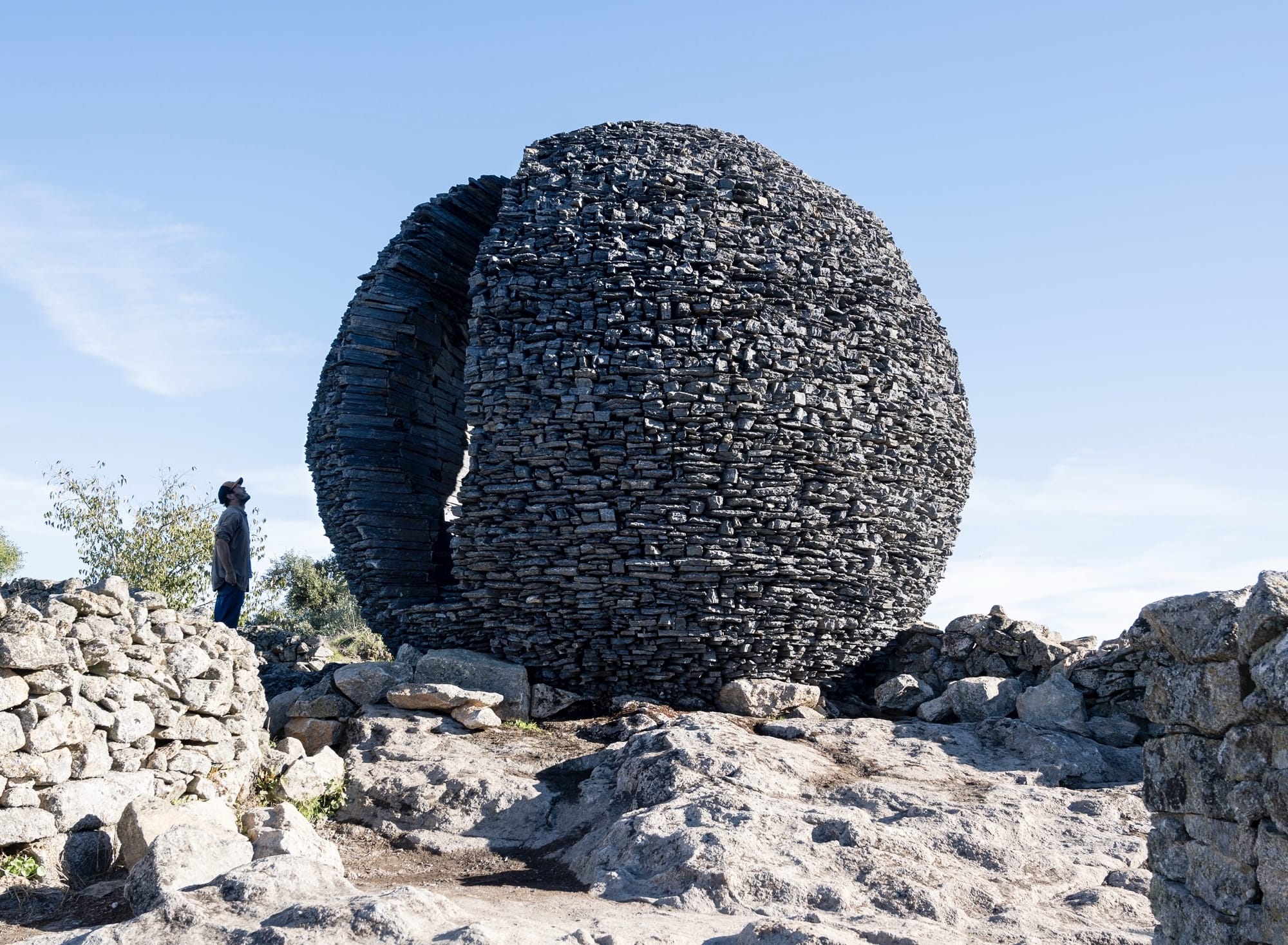 A sculptural installation outdoors made of black schist in the shape of a sphere with apertures for people to step into and see out of